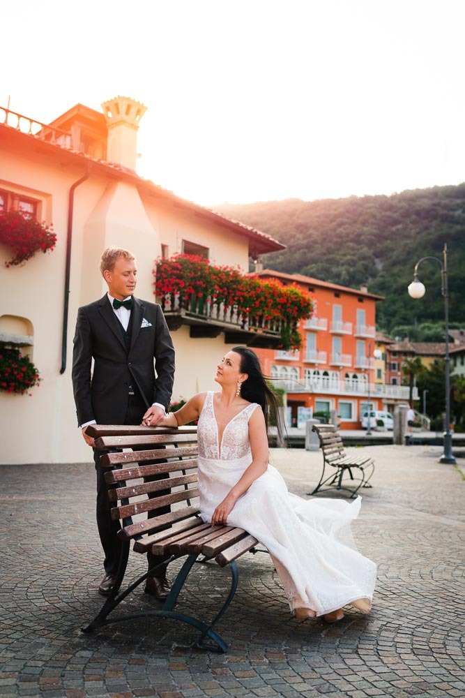 Para młoda w eleganckich strojach pozuje na drewnianej ławce na tle malowniczego miasteczka z budynkami ozdobionymi kwiatami i górami w oddali.