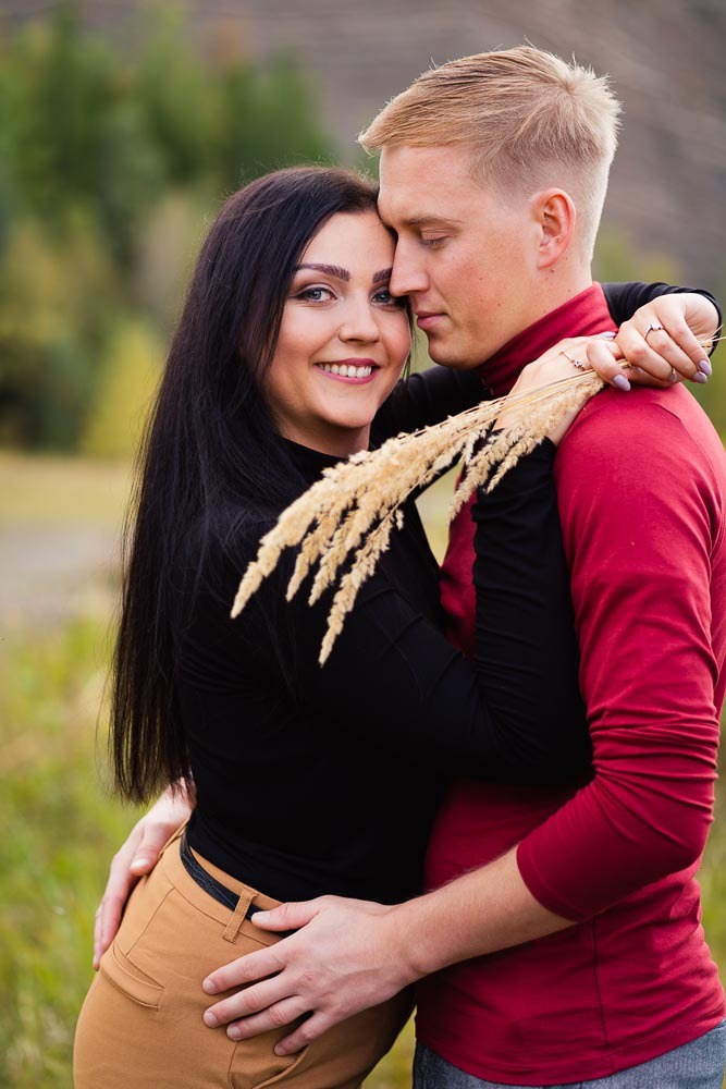 Uśmiechnięta brunetka w czarnej bluzce i brązowych spodniach, wtulona w blondyna w czerwonym golfie, trzymająca w dłoniach pęk suchych traw na tle zieleni.