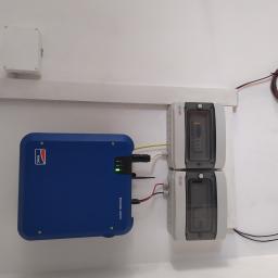 Solarwat Jacek Chancewicz - Niebieski falownik SMA Sunny Tripower zamontowany na białej ścianie, obok dwa szare zabezpieczenia ETI z widocznymi wewnątrz bezpiecznikami, całość połączona przewodami.