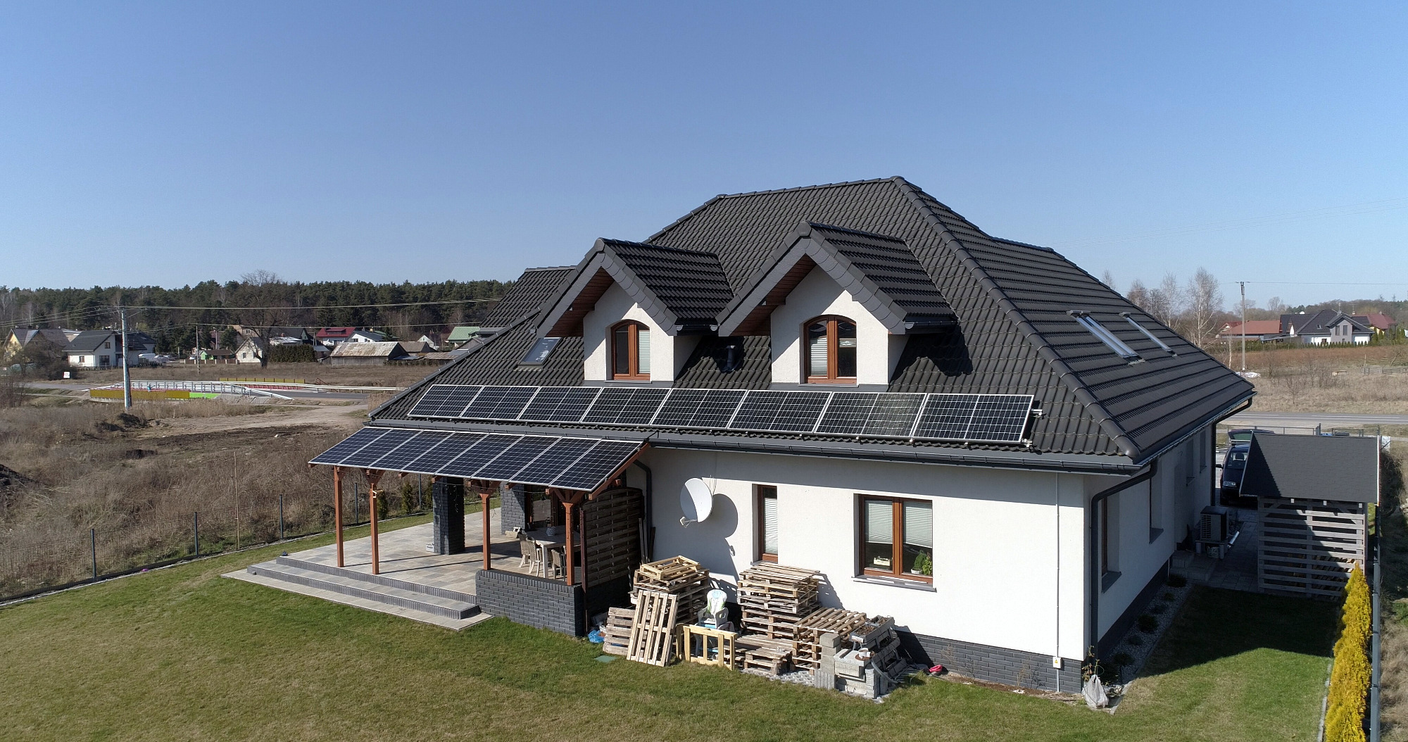 Dom jednorodzinny z panelami słonecznymi na dachu i nad tarasem, widok z góry, palety drewniane składowane obok budynku.