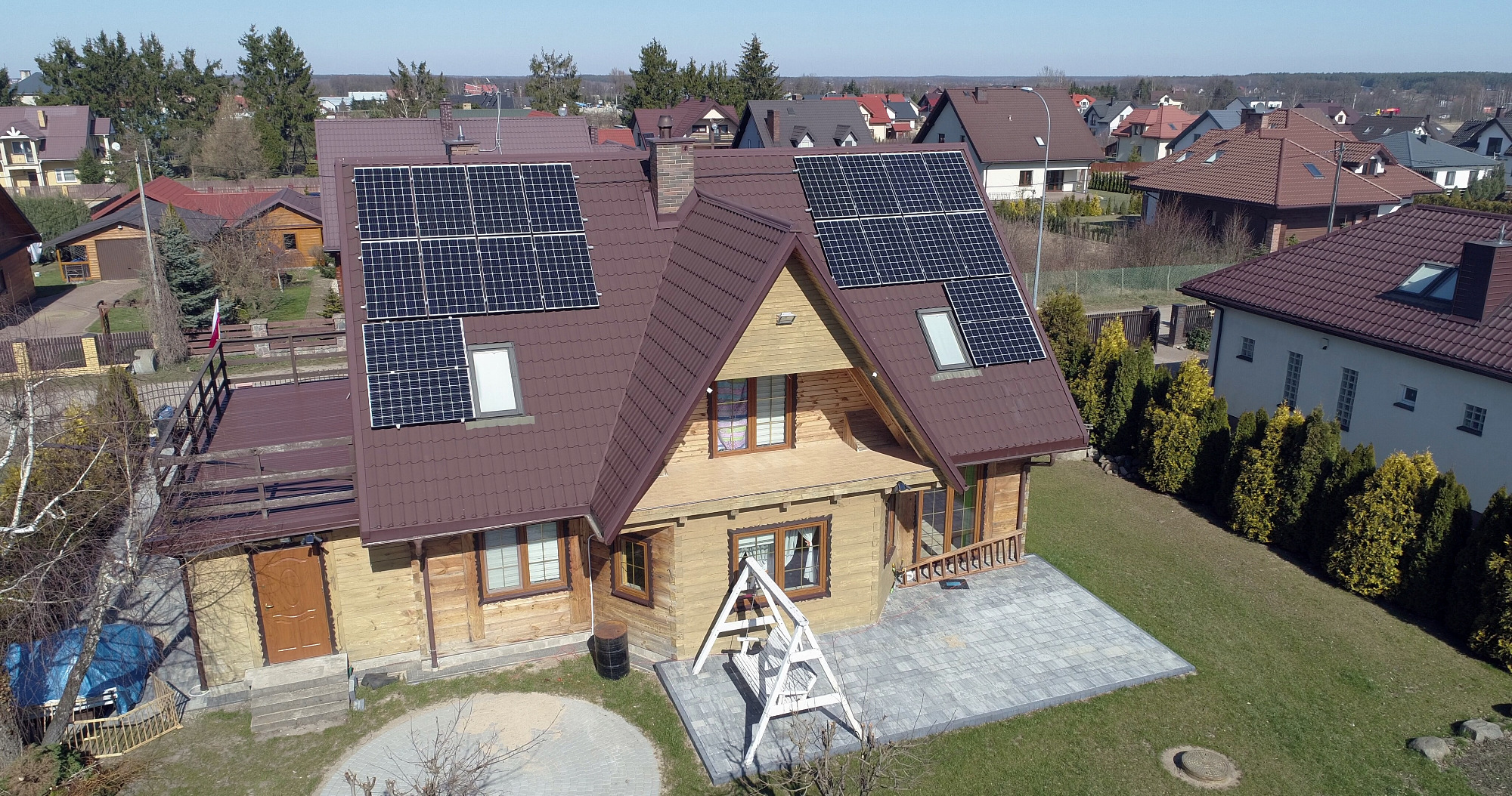 Drewniany dom jednorodzinny z brązowym dachem, na którym zamontowane są panele słoneczne; widok z lotu ptaka na osiedle domów.