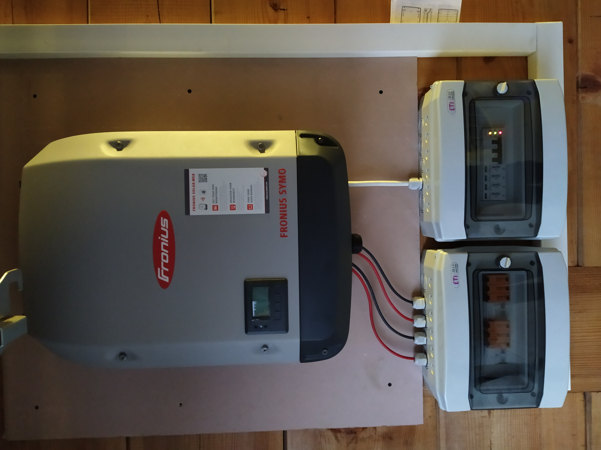 Szary inwerter Fronius Symo zamontowany na płycie z podłączonymi kablami do dwóch białych skrzynek ETI SIMTEC z widocznymi bezpiecznikami wewnątrz, całość na tle boazerii.