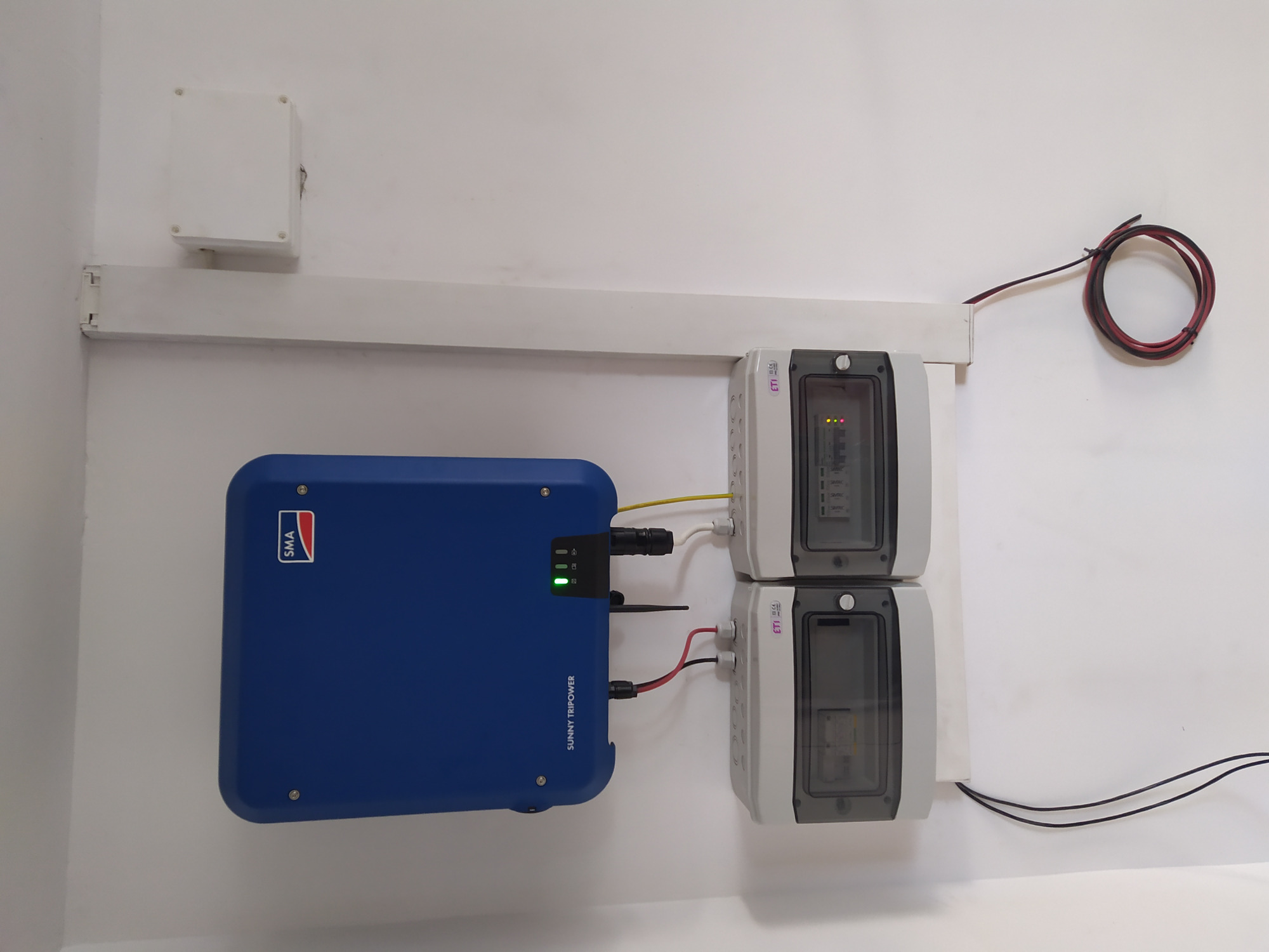 Niebieski falownik SMA Sunny Tripower zamontowany na białej ścianie, obok dwa szare zabezpieczenia ETI z widocznymi wewnątrz bezpiecznikami, całość połączona przewodami.