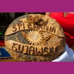 Okrągły, dekoracyjny bochenek chleba z napisem 'SPIŻARNIA KUJAWSKA', ozdobiony kwiatami z ciasta i posypany różnymi ziarnami.