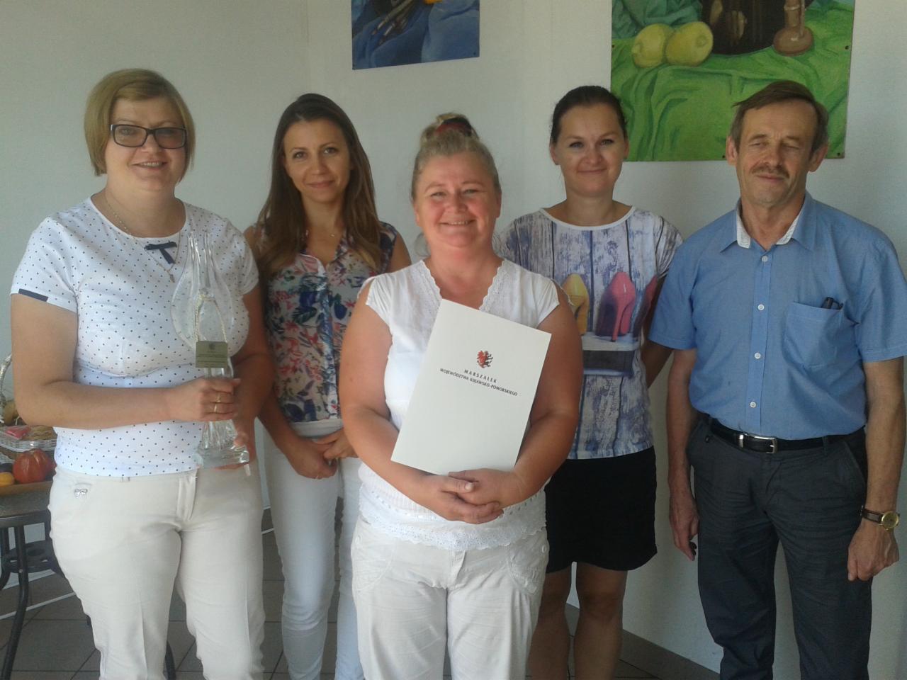 Grupa pięciu osób, w tym kobieta trzymająca certyfikat Marszałka Województwa Kujawsko-Pomorskiego oraz kobieta z przezroczystą statuetką, stoją na tle ściany z obrazami.