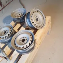 Cztery surowe, stalowe felgi samochodowe, leżące na drewnianej palecie, prawdopodobnie po procesie oczyszczania, z widocznym przewodem i ścianą w tle.