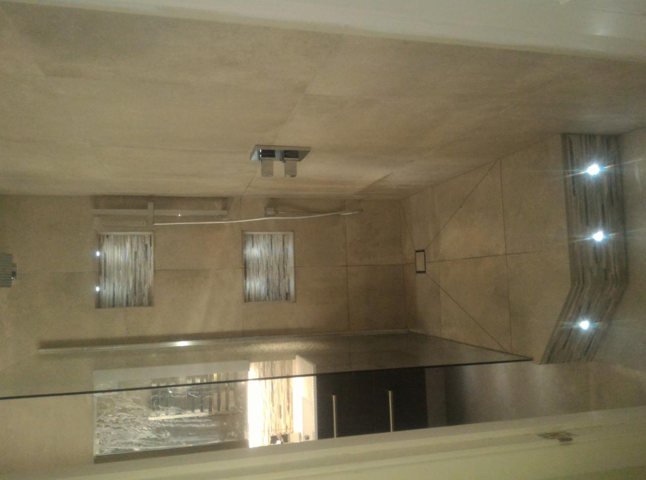 Wykończona łazienka z beżowymi płytkami, szklaną kabiną prysznicową i dekoracyjnym oświetleniem ściennym z trzema punktami świetlnymi.