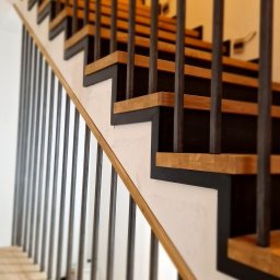 EBM Stolarstwo Tradycyjne - Nowoczesne schody z drewnianymi stopniami, czarnymi podstopnicami i metalową balustradą z pionowych prętów, widok z dołu w górę.