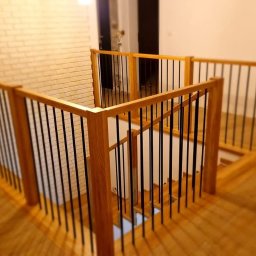 EBM Stolarstwo Tradycyjne - Drewniana balustrada schodowa z czarnymi, pionowymi prętami, widok z góry na schody z drewnianymi stopniami i ceglaną ścianą w tle.