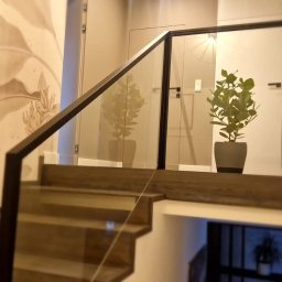 EBM Stolarstwo Tradycyjne - Szklana balustrada schodowa z ciemną, drewnianą ramą i poręczą, z rośliną w doniczce na podeście, na tle ściany z motywem roślinnym.