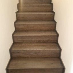 EBM Stolarstwo Tradycyjne - Proste, drewniane schody wewnętrzne z ciemnego drewna, widok z dołu, oświetlone naturalnym światłem wpadającym z góry.