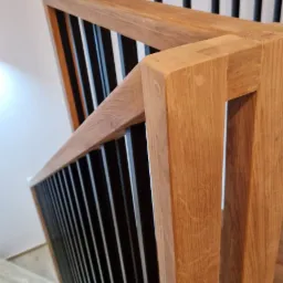 Fragment dębowej balustrady schodowej z czarnymi, metalowymi tralkami, widok z góry na jasne stopnie schodów.