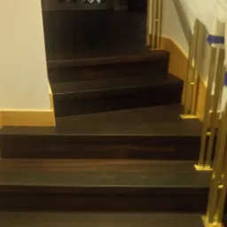 Ciemne stopnie schodów z metalową, złotą balustradą zabezpieczoną folią, widok z dołu na okno w tle.