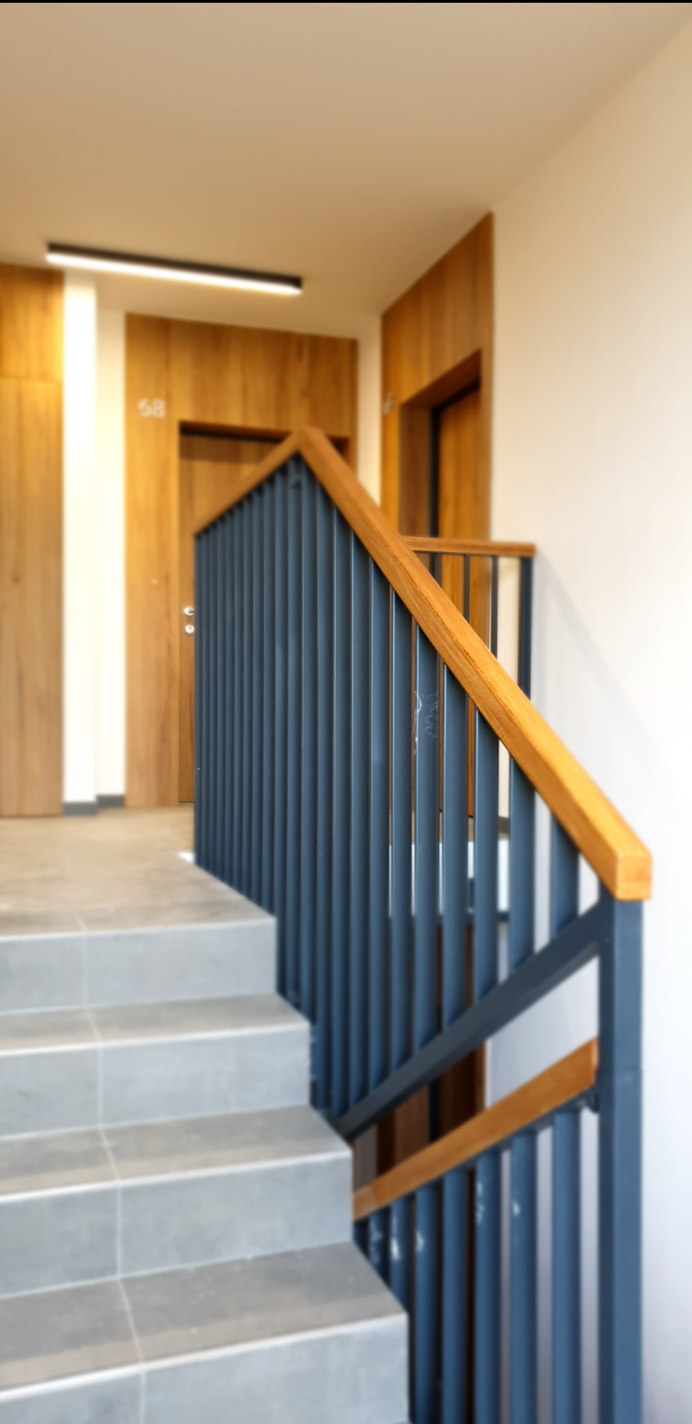 Szare schody z metalową balustradą w kolorze grafitowym i drewnianą poręczą, widok z dołu, drzwi z numerem mieszkania w tle.