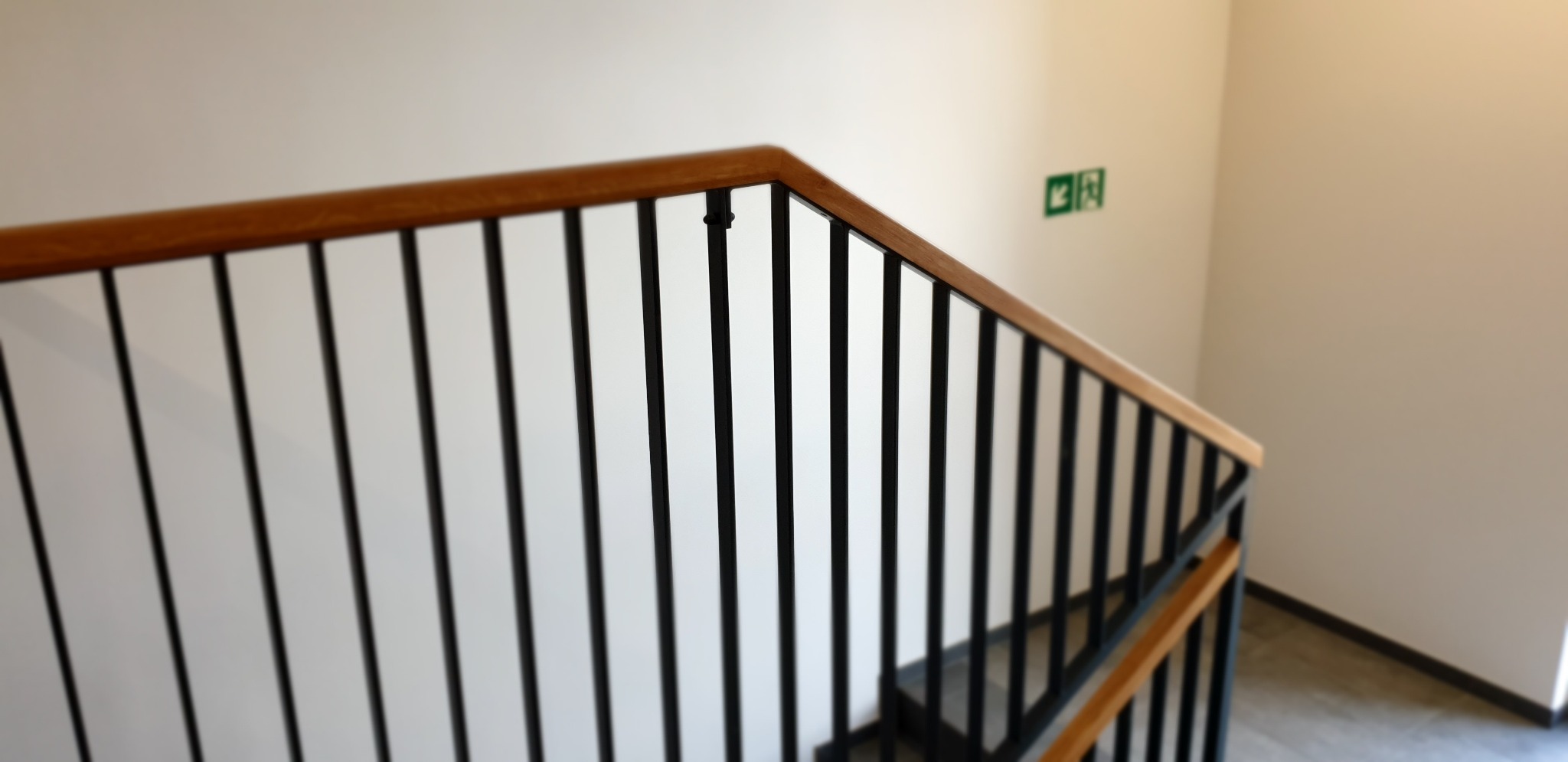 Nowoczesna balustrada schodowa z drewnianą poręczą i czarnymi, metalowymi słupkami, widok z góry na bieg schodów z betonowymi stopniami, znak ewakuacyjny na ścianie.