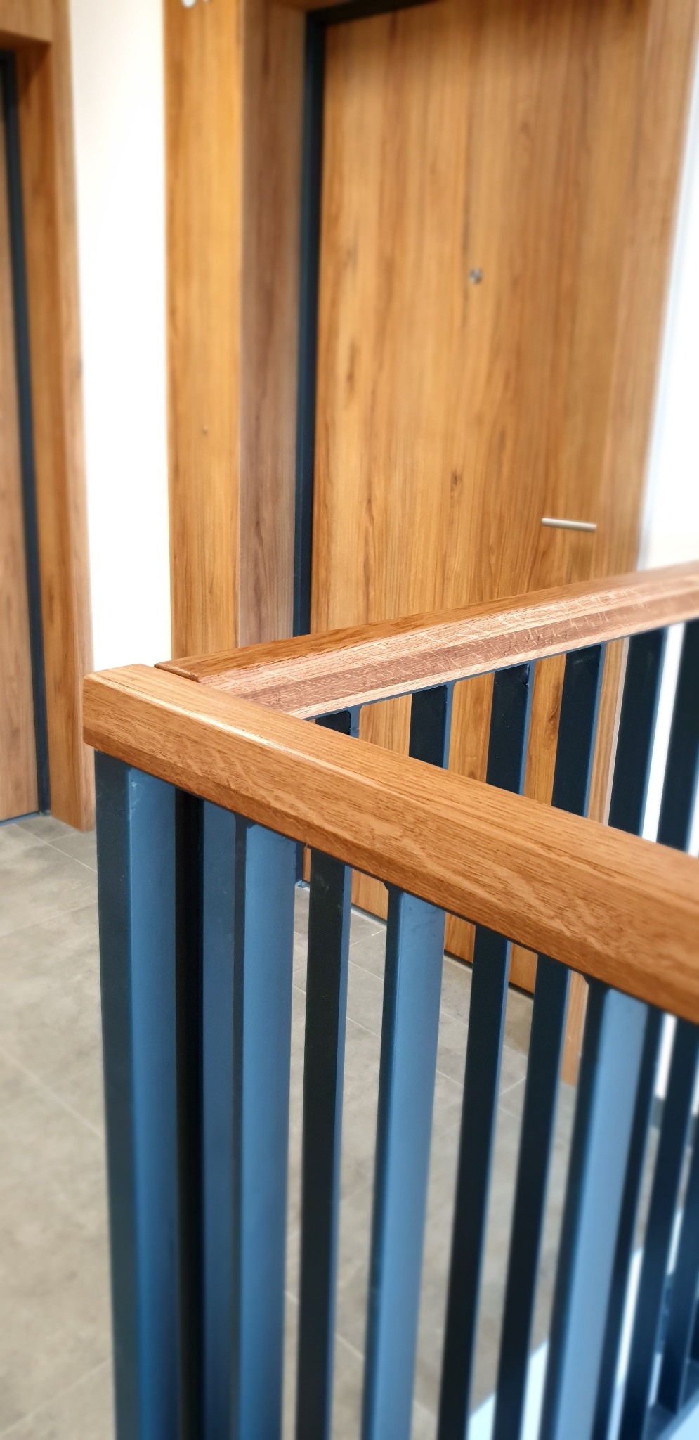 Fragment drewniano-metalowej balustrady schodowej z drewnianym pochwytem i pionowymi, metalowymi słupkami w kolorze grafitowym, na tle drewnianych drzwi.