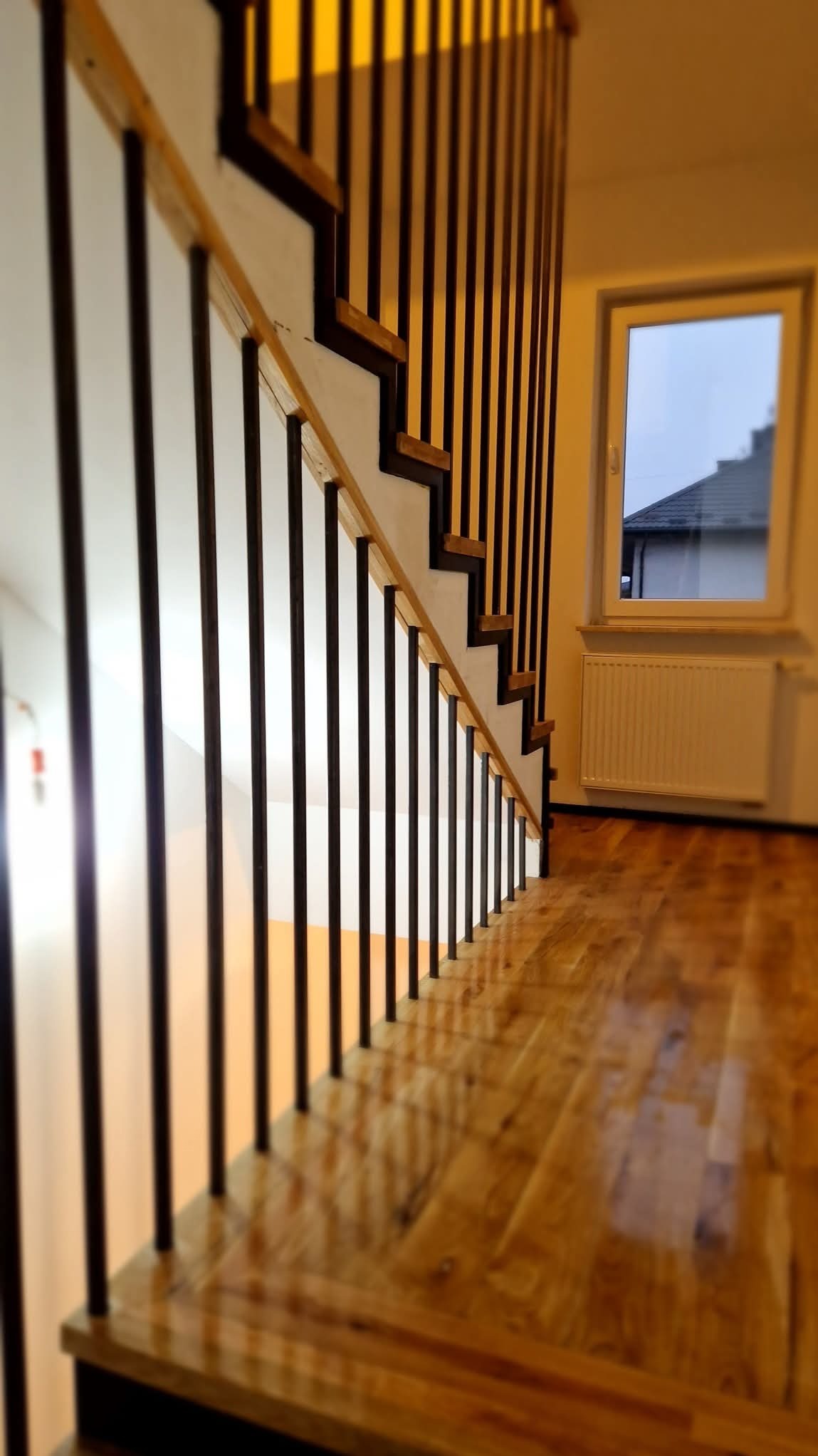 Nowoczesne schody z drewnianymi stopniami i czarnymi, metalowymi balustradami, widok z dołu, polerowana drewniana podłoga, okno w tle.
