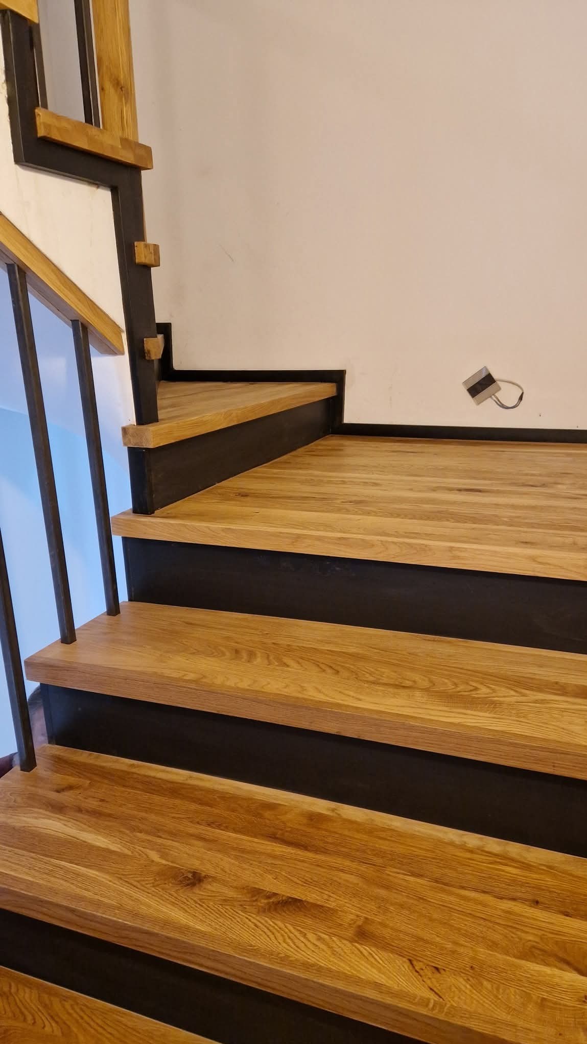 Fragment nowoczesnych schodów z drewnianymi stopniami o ciepłym odcieniu i czarną konstrukcją nośną oraz metalową balustradą, widoczne niedokończone podłączenie elektryczne na ścianie.