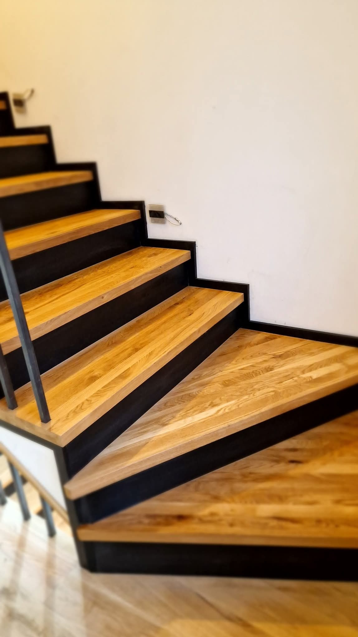 Nowoczesne schody z drewnianymi stopniami i czarną metalową konstrukcją, widoczne z dołu, z minimalistyczną białą ścianą w tle.