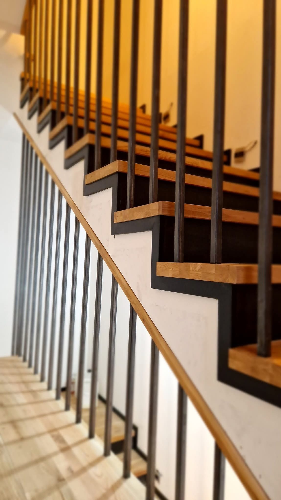 Nowoczesne schody z drewnianymi stopniami, czarnymi podstopnicami i metalową balustradą z pionowych prętów, widok z dołu w górę.