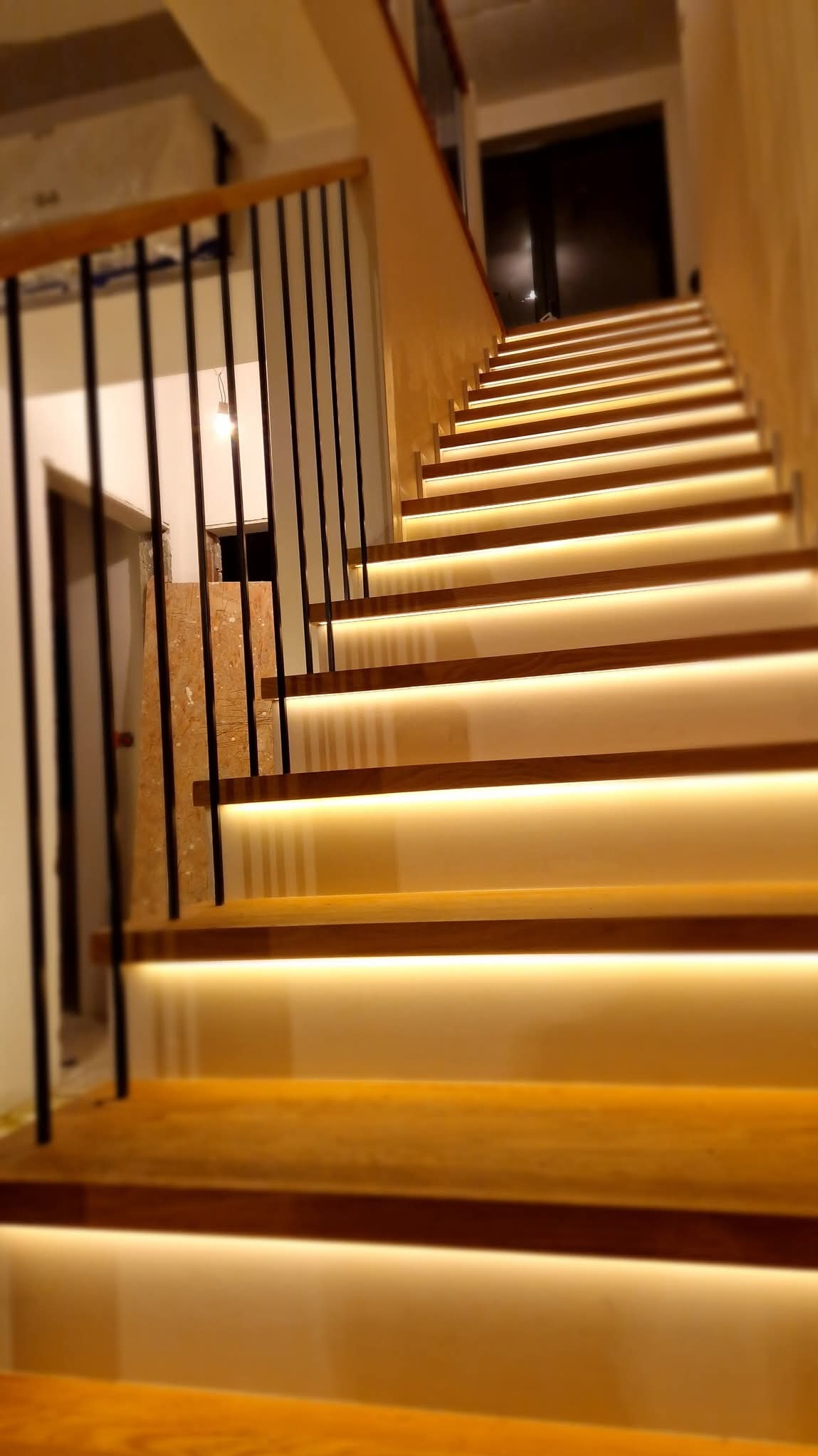 Nowoczesne schody z drewnianymi stopniami podświetlonymi ciepłym światłem LED, czarne metalowe barierki, widok z dołu ku górze.