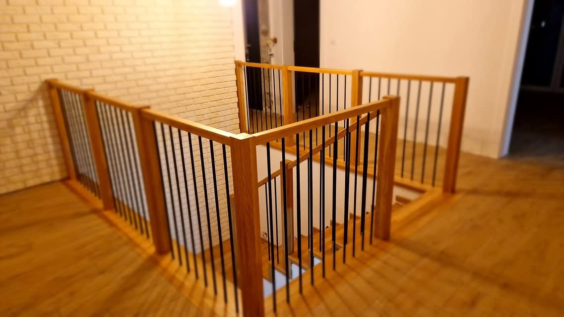 Drewniana balustrada schodowa z czarnymi, pionowymi prętami, widok z góry na schody z drewnianymi stopniami i ceglaną ścianą w tle.