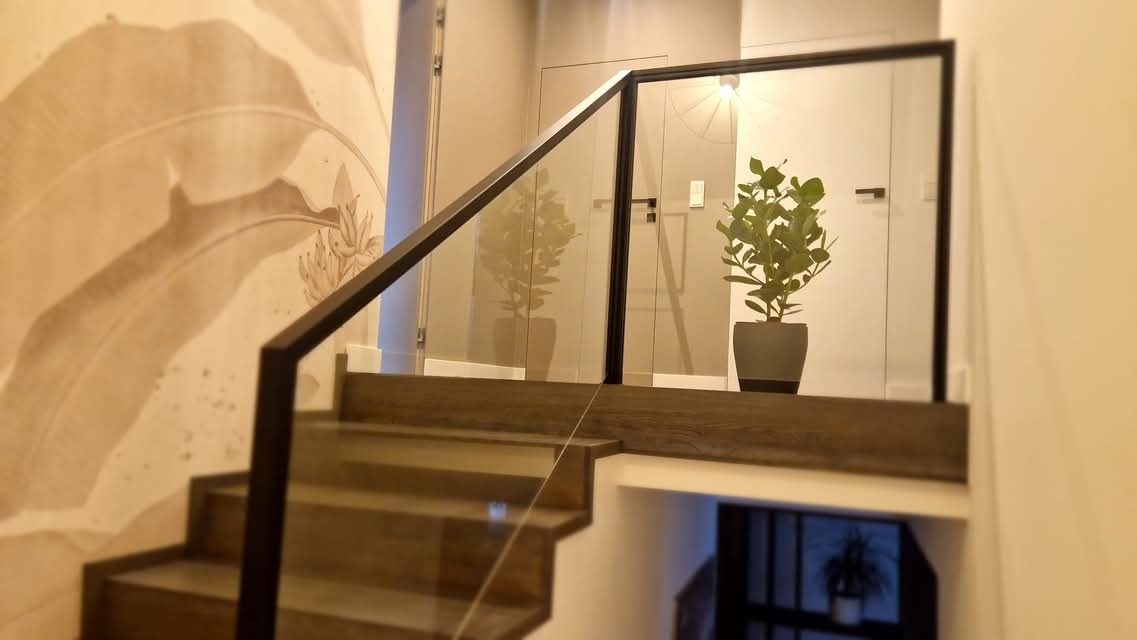 Szklana balustrada schodowa z ciemną, drewnianą ramą i poręczą, z rośliną w doniczce na podeście, na tle ściany z motywem roślinnym.