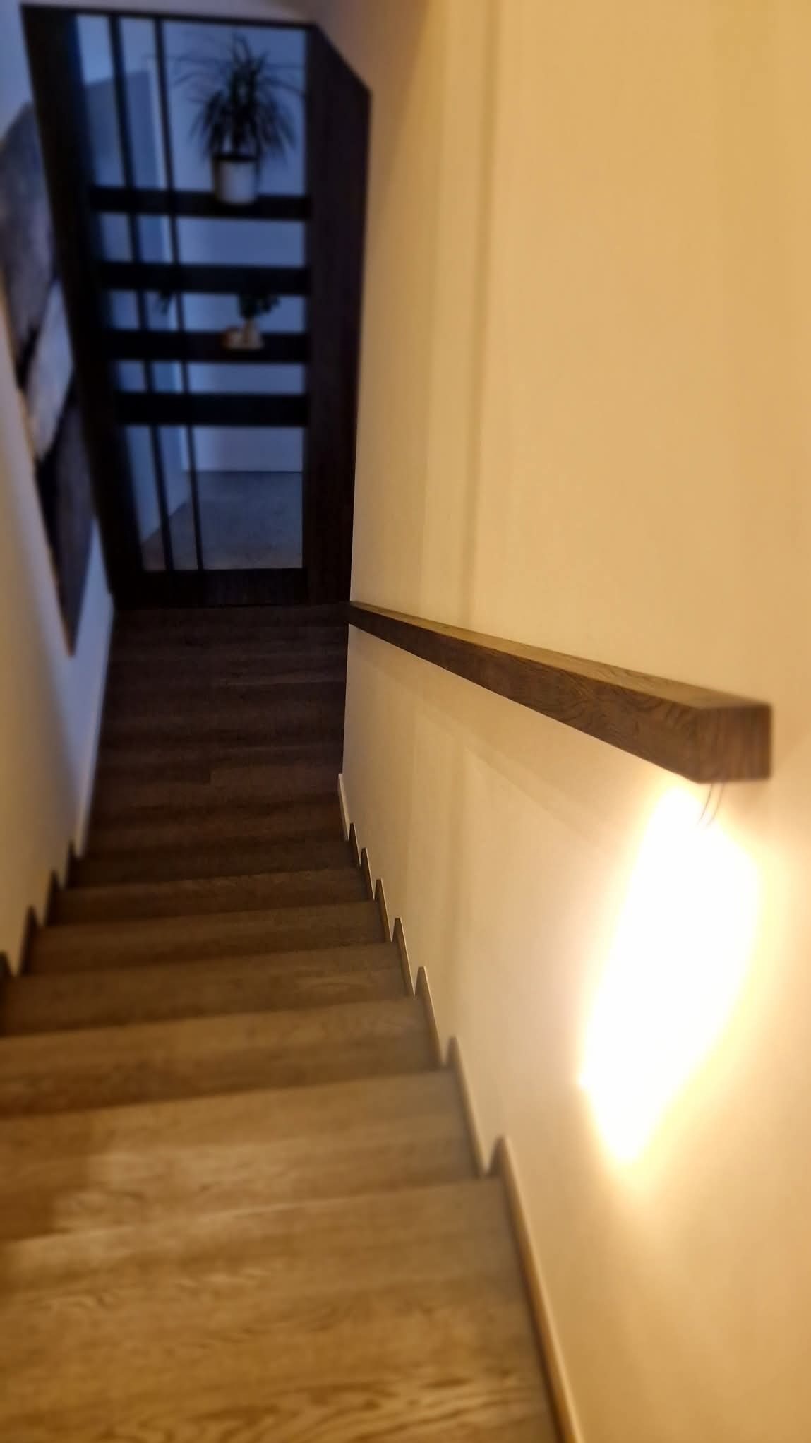 Widok z góry na jasne drewniane schody z minimalistyczną poręczą zamontowaną na ścianie, oświetlone ciepłym światłem.