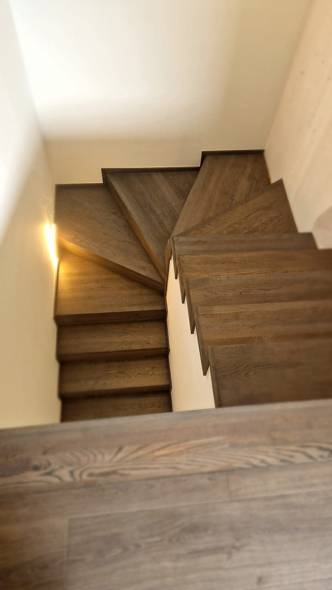 Kątowy widok z góry na brązowe, drewniane schody z zabiegiem, z widocznym usłojeniem drewna i subtelnym oświetleniem od strony ściany.