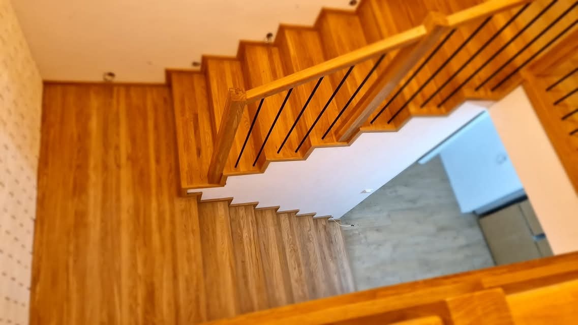 Widok z góry na jasne, drewniane schody z czarnymi, metalowymi prętami w balustradzie, dopasowane kolorystycznie do drewnianej boazerii ściennej.