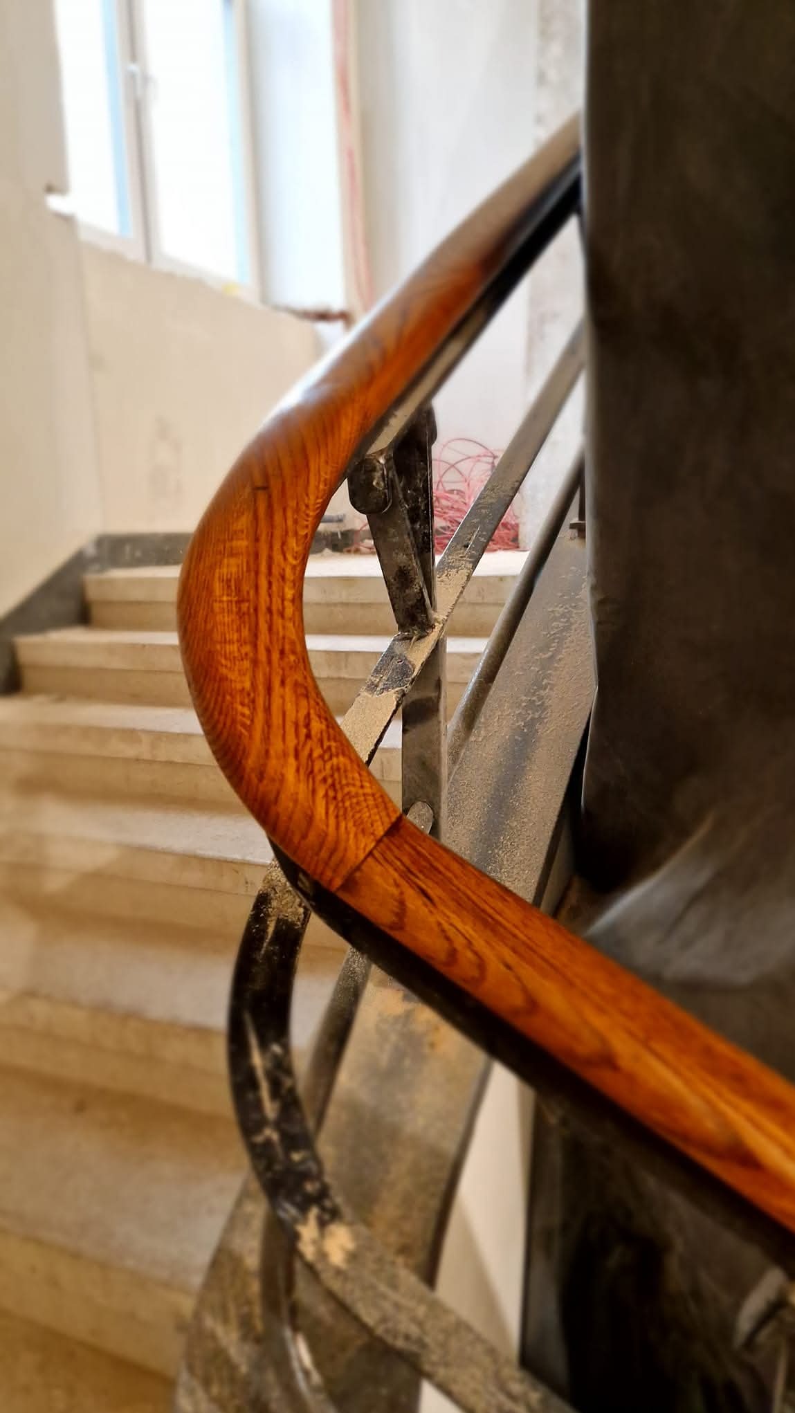 Fragment drewnianej poręczy schodowej z metalowymi elementami konstrukcyjnymi, widoczne ślady pyłu po obróbce, w tle schody betonowe i okno.