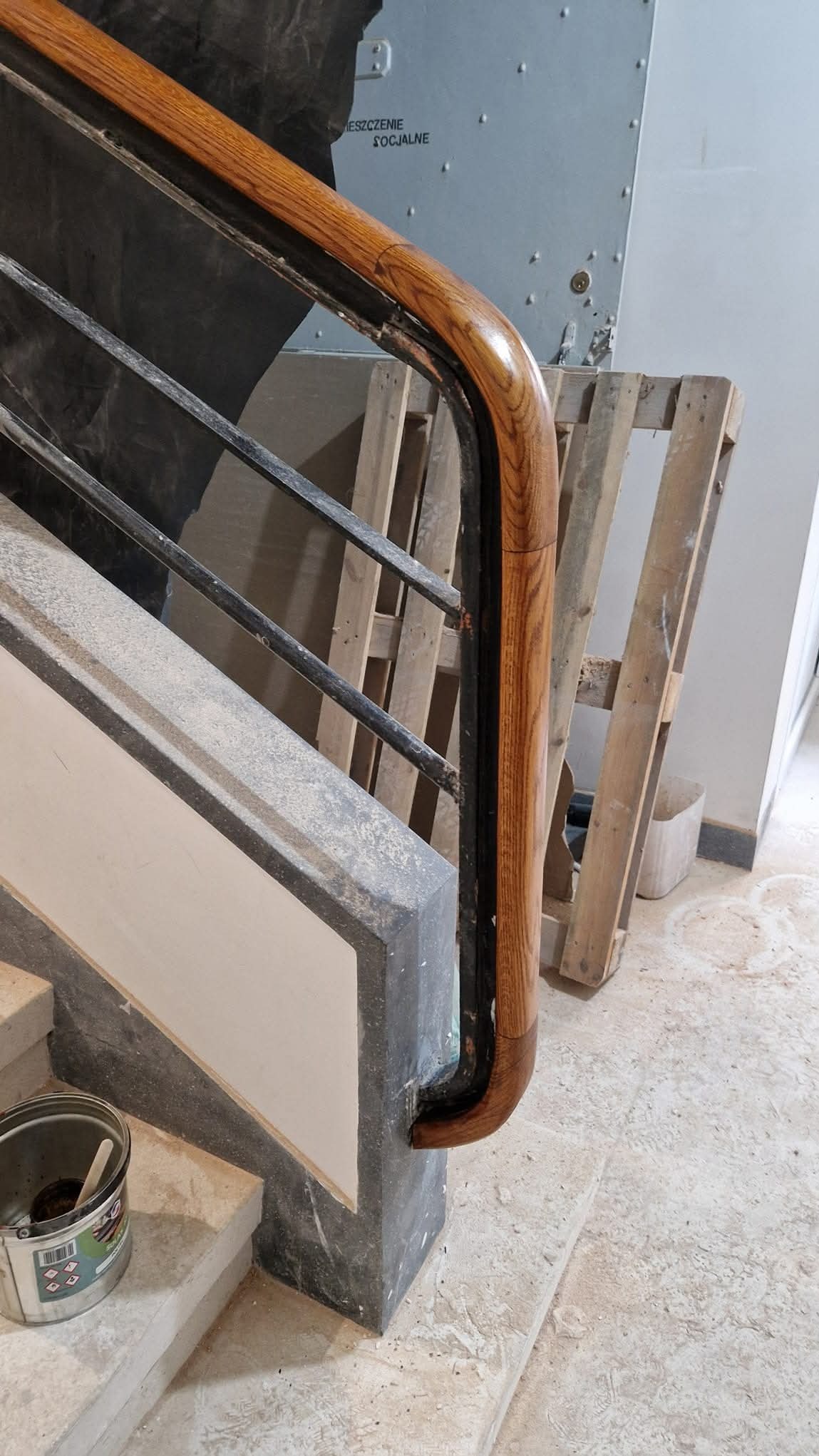 Fragment klatki schodowej w trakcie renowacji: drewniana poręcz z metalowymi elementami, widoczne ślady prac remontowych, puszka z farbą na schodach, w tle palety.