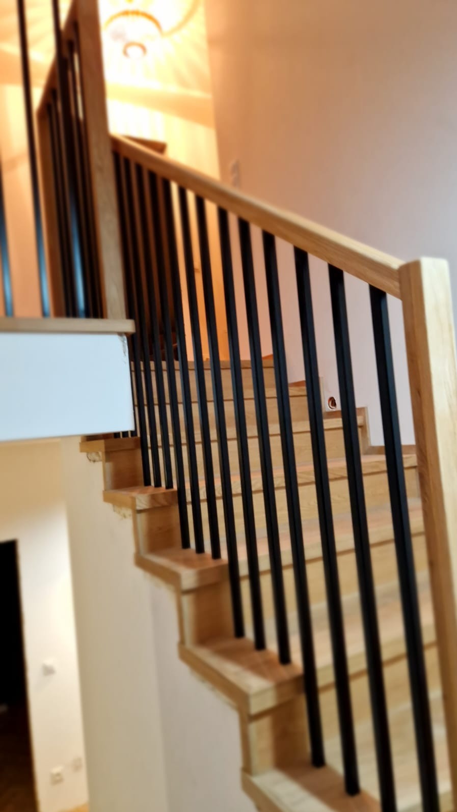 Jasne, drewniane schody z czarnymi, pionowymi balustradami, widok z dołu, oświetlenie sufitowe w tle.