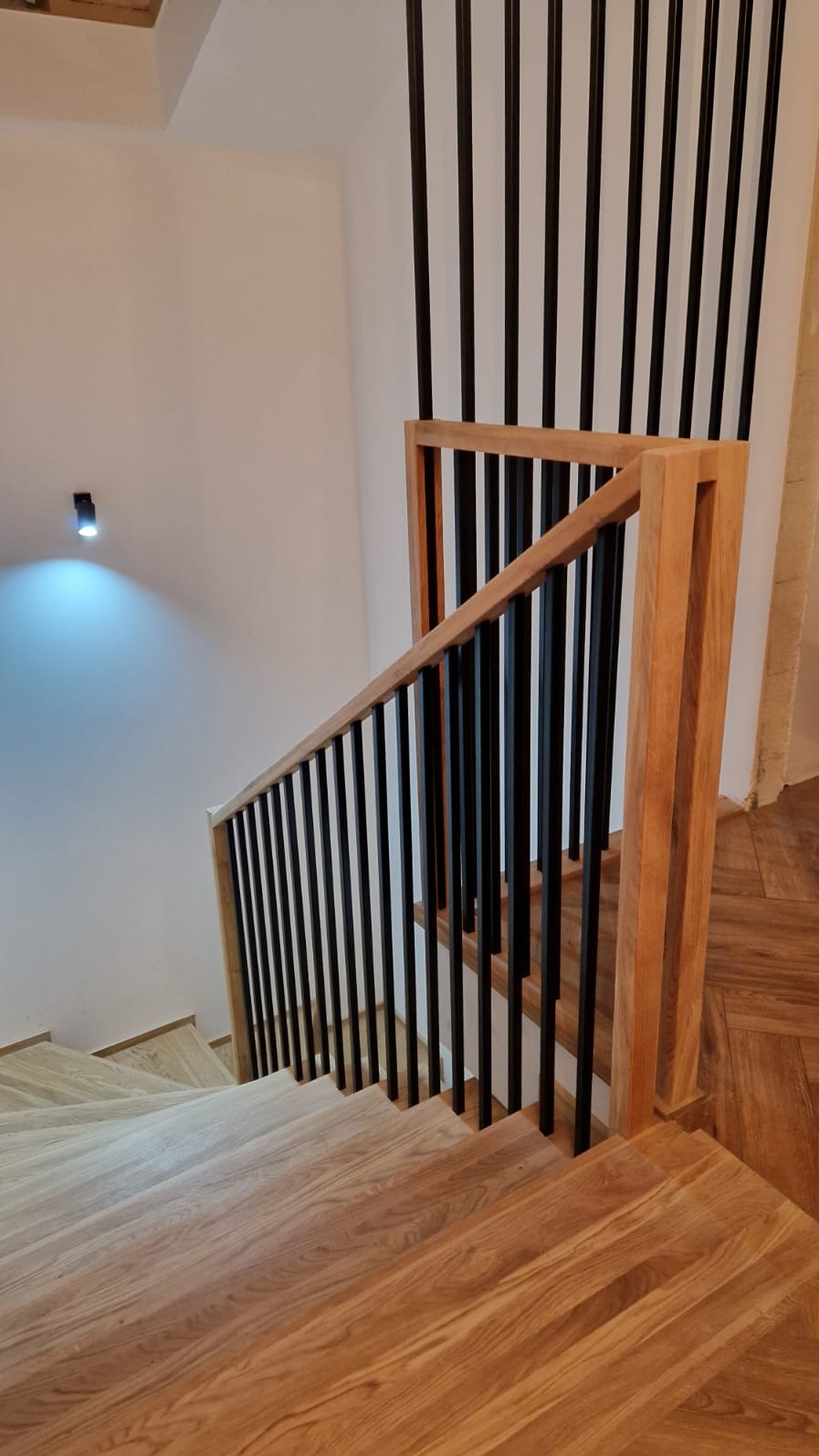 Nowoczesne schody drewniane z balustradą z czarnych, pionowych prętów i drewnianą poręczą, oświetlone punktowym światłem na białej ścianie.