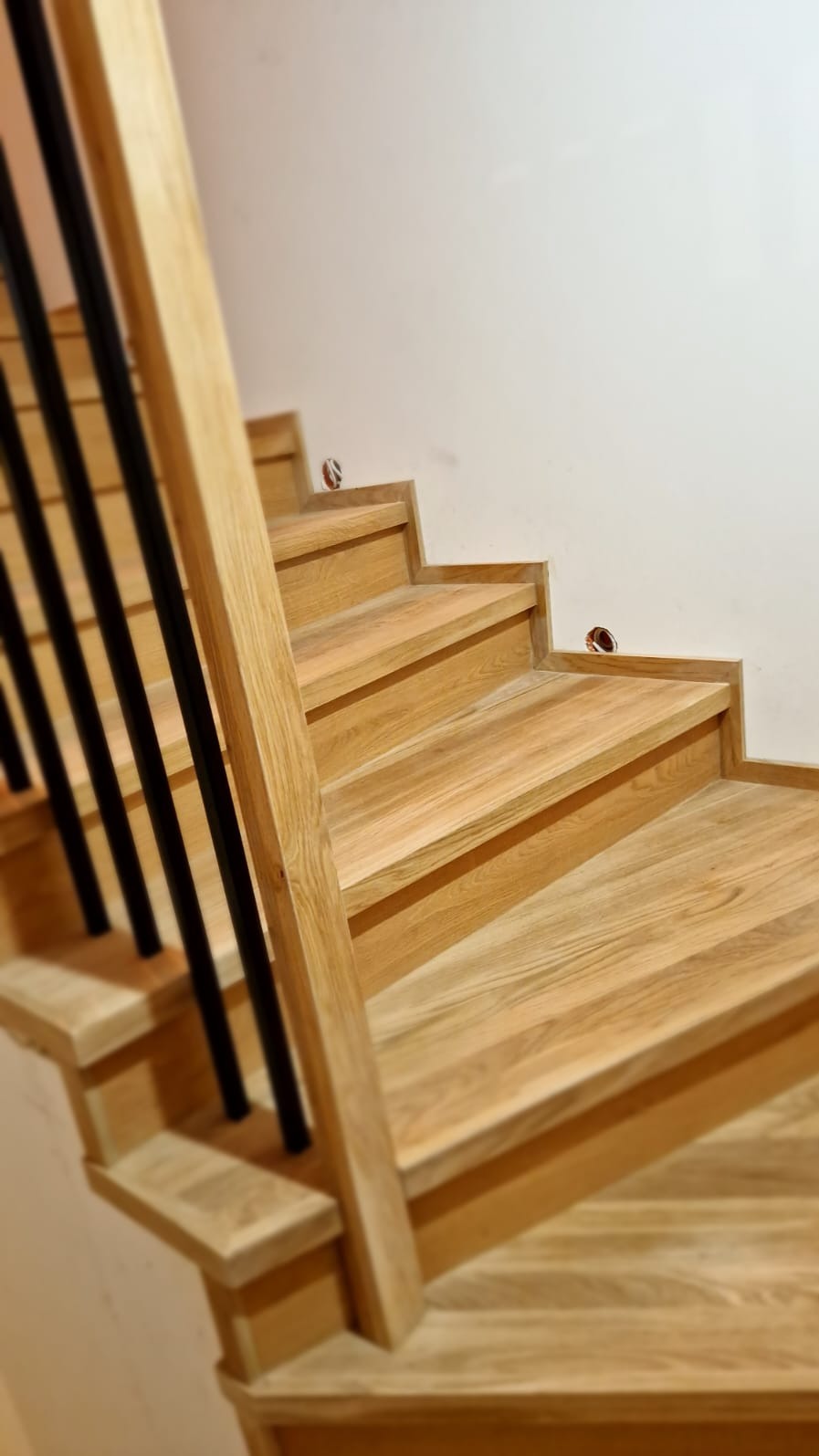 Jasne, dębowe stopnie schodów z czarnymi, pionowymi elementami balustrady, widoczne otwory instalacyjne na ścianie.