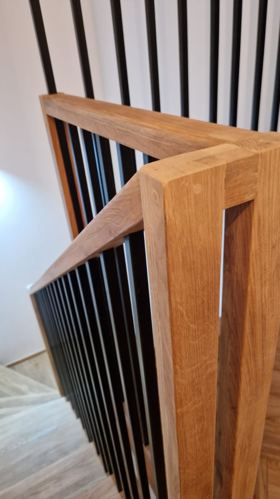 Fragment dębowej balustrady schodowej z czarnymi, metalowymi tralkami, widok z góry na jasne stopnie schodów.
