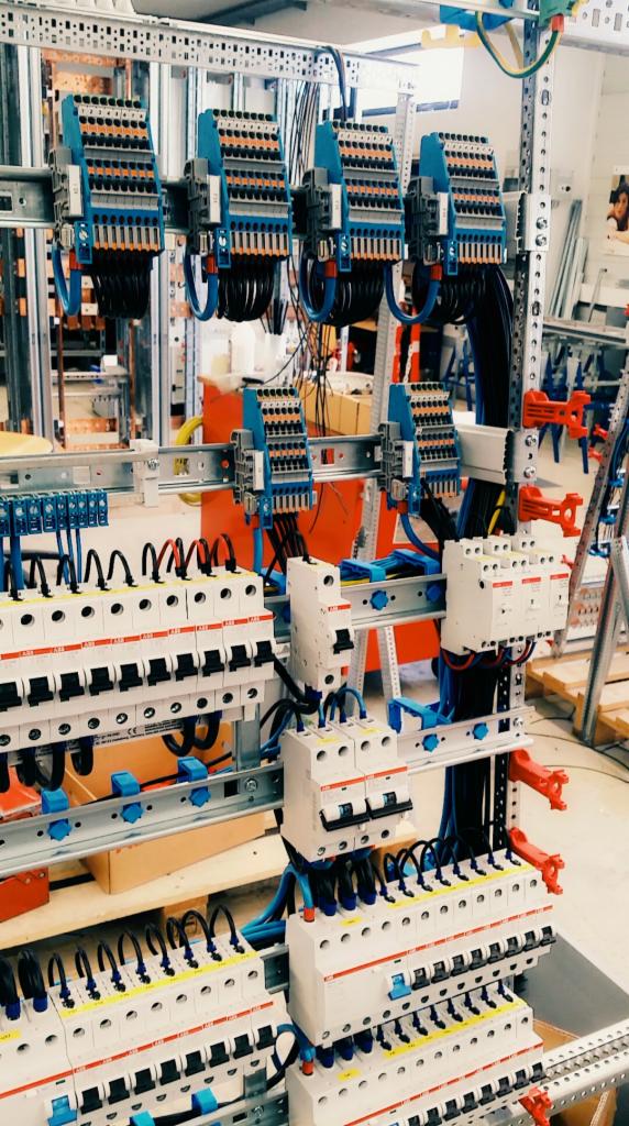 Rozdzielnica elektryczna z zabezpieczeniami nadprądowymi i listwami zaciskowymi, widoczne przewody w kolorach niebieskim i czarnym, montaż na szynie DIN, tło warsztatowe.
