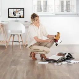 Kobieta w kucki przegląda próbki materiałów wykończeniowych na podłodze z paneli w jasnym wnętrzu, z biurkiem i komputerem w tle.