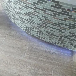 Zaokrąglona obudowa wanny wykończona mozaiką z płytek w odcieniach szarości i beżu, przylegająca do podłogi wyłożonej panelami imitującymi drewno.