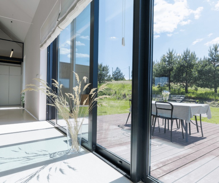 Nowoczesne drzwi przesuwne z czarnymi ramami, widok na taras z drewnianą podłogą i stół nakryty obrusem w kratę, w wazonie trawy ozdobne.