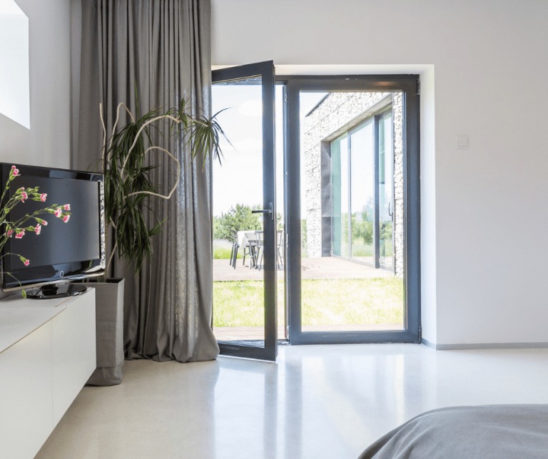 Nowoczesne, szare drzwi balkonowe uchylone do wewnątrz, widok na taras z meblami ogrodowymi i trawnik, wnętrze z telewizorem i szarą zasłoną.