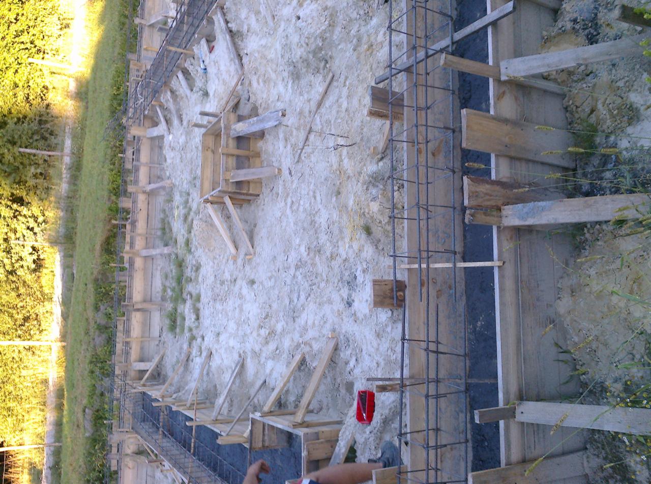 Wykop pod fundamenty z szalunkiem drewnianym i zbrojeniem stalowym, widoczny czerwony pojemnik na narzędzia, trawa w tle.