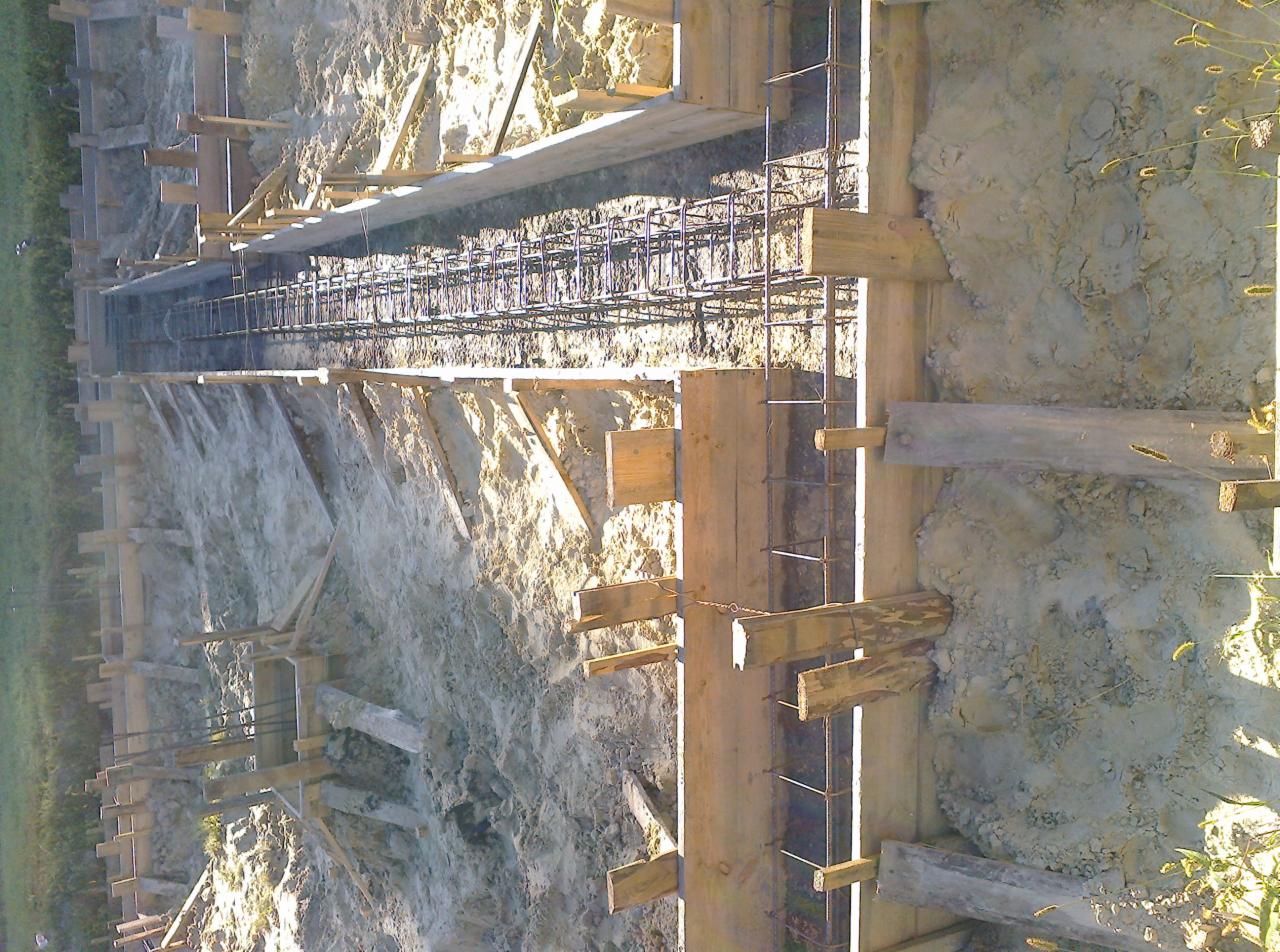 Szczegółowe ujęcie szalunku i zbrojenia fundamentu w wykopie, przygotowanego do zalania betonem, z widocznymi drewnianymi elementami konstrukcyjnymi i stalowymi prętami zbrojeniowymi.