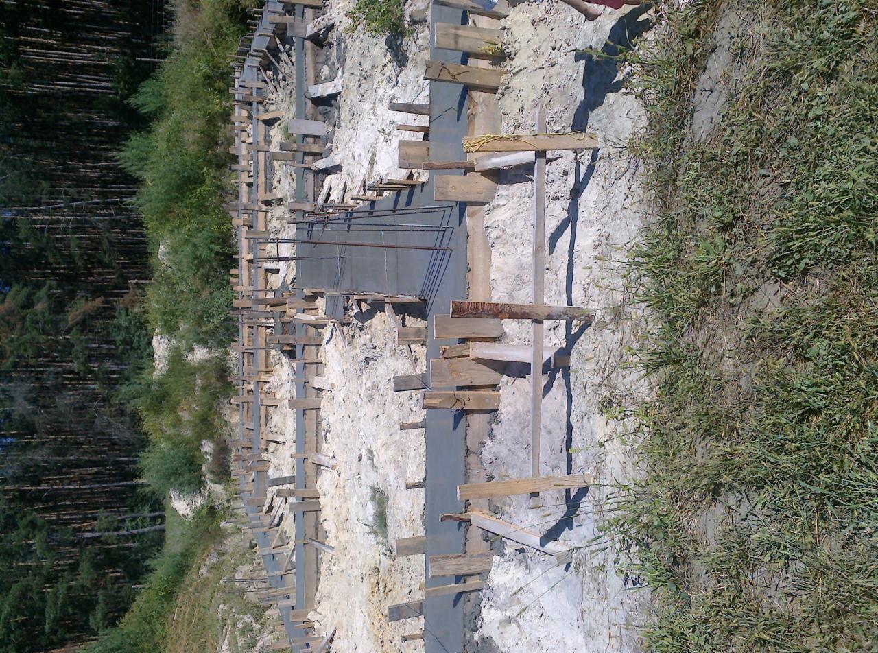 Wykop pod fundamenty z drewnianym szalunkiem i częściowo wylanym betonem, widoczne zbrojenia, teren budowy otoczony zielenią.