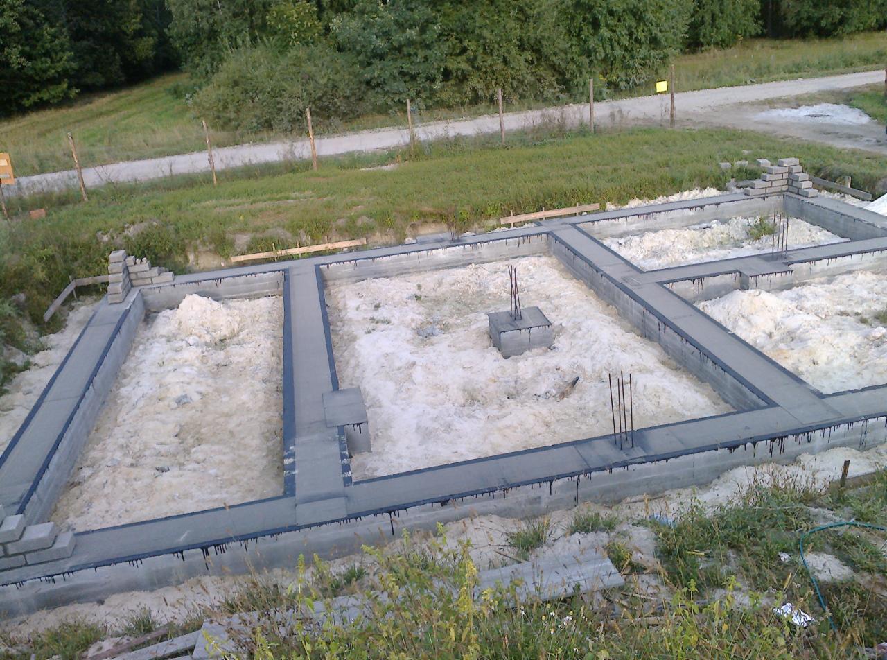 Zalane betonem fundamenty z wystającymi prętami zbrojeniowymi, widok z góry na plac budowy w otoczeniu zieleni.