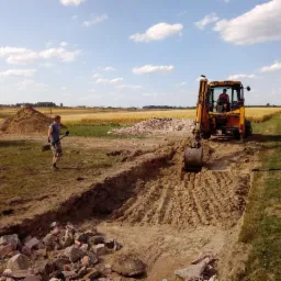 Żółta koparko-ładowarka podczas tworzenia wykopu na polu, pracownik z łopatą obok, sterta gruzu w tle, pole zboża w oddali.