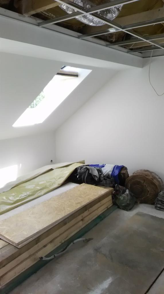 Poddasze w trakcie remontu z oknem dachowym, przygotowane płyty OSB, folia ochronna i materiały izolacyjne pod sufitem z widoczną konstrukcją metalową