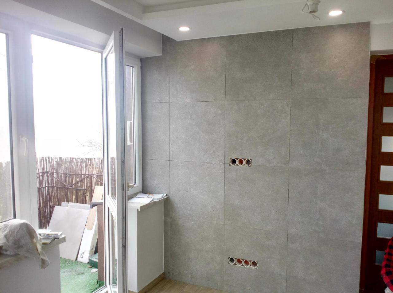 Szara ściana wyłożona płytkami w trakcie montażu, widoczne puszki elektryczne bez osprzętu, otwarte okno balkonowe, podłoga wyłożona panelami.