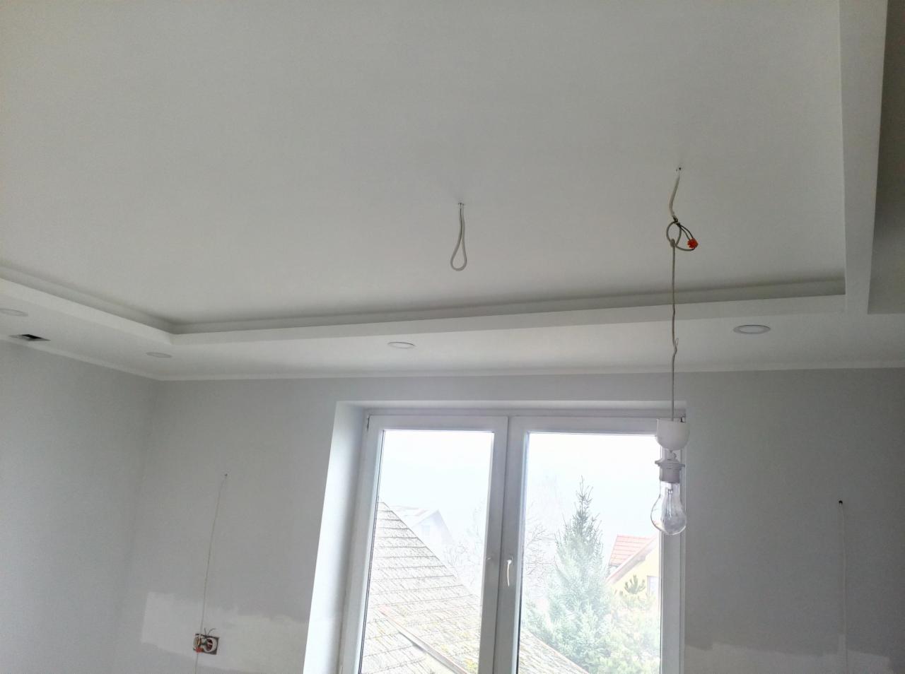 Białe wnętrze w trakcie prac wykończeniowych: widoczne gładzie na ścianach, sufit podwieszany z oświetleniem punktowym, okno z widokiem na zewnątrz, oraz prowizoryczne oświetlenie w postaci...