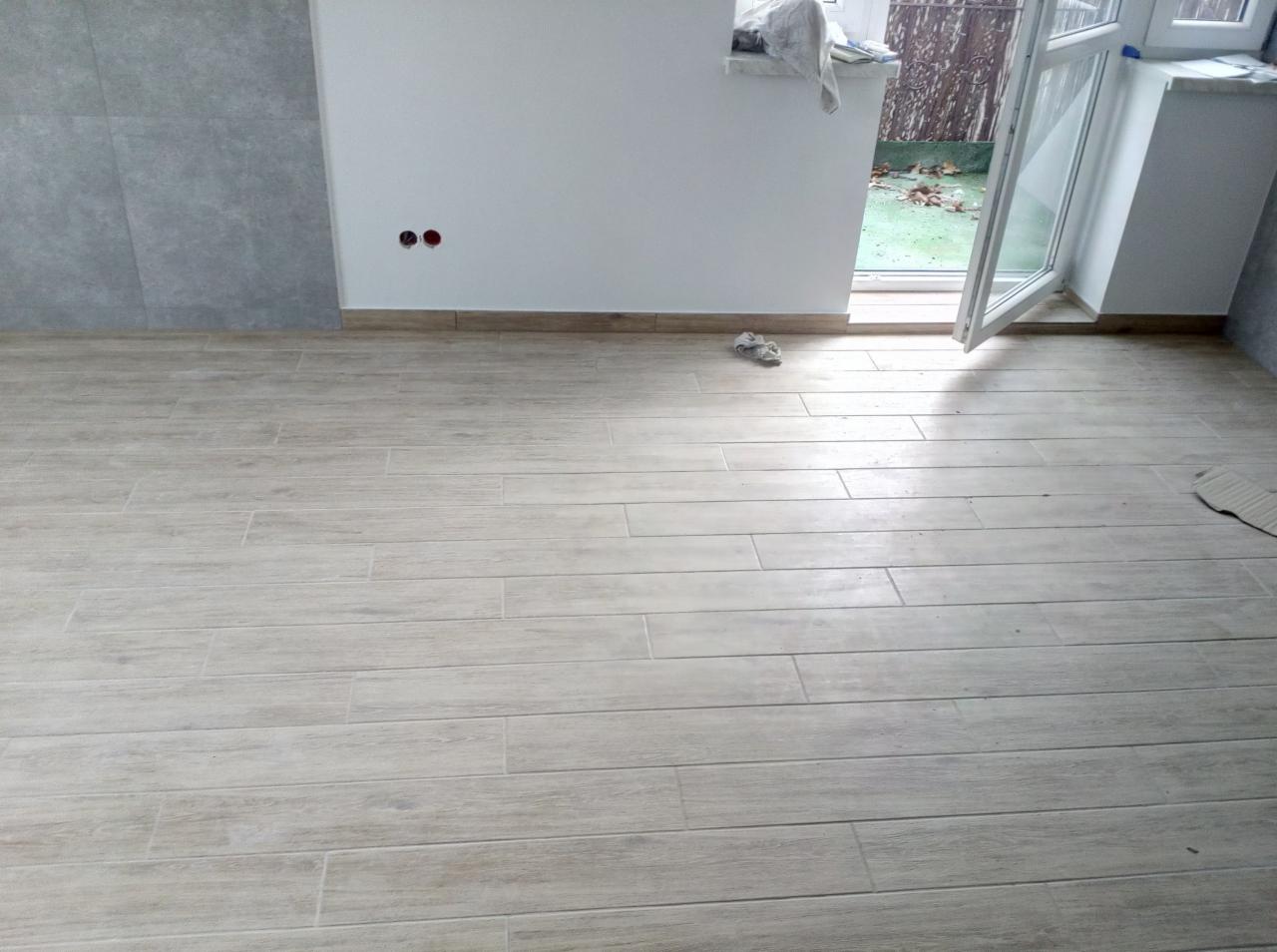 Jasne, drewnopodobne płytki podłogowe ułożone w nowym pomieszczeniu z otwartym oknem na ogród, widoczne gniazdka elektryczne bez osprzętu.